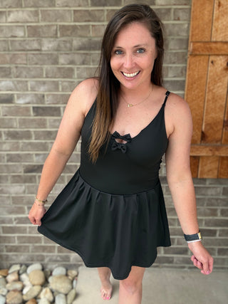 A woman modeling a black sleeveless swim dress with a ruffled peplum skirt and a small bow detail at the neckline, standing in front of a gray brick wall.