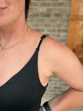 Close-up of a woman wearing a black one-shoulder tank top, showing the shoulder strap and side neckline, with a brick wall background.