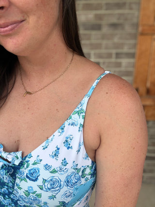 A close-up lifestyle shot of a woman wearing a delicate gold necklace with a small leaf pendant, paired with a white spaghetti-strap top featuring a blue and green floral print.
