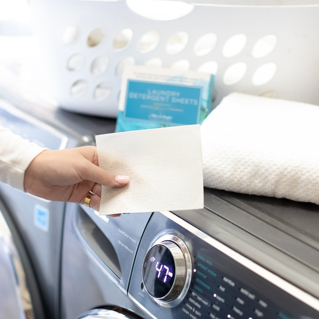 Laundry Detergent Sheets in a blue box scented with Mixologie's Boujee Fragrance. Eco-friendly, dry-free, dissolvable sheets provide superior cleaning power with a sophisticated, fresh fragrance! Detergent sheets box has 30 sheets dimensions are 4.375 in X 5.5 inches. Sheet in model hand in front of washing machine